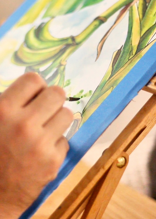 A close-up of a hand painting a green and yellow leaf illustration on a canvas mounted on a wooden easel.