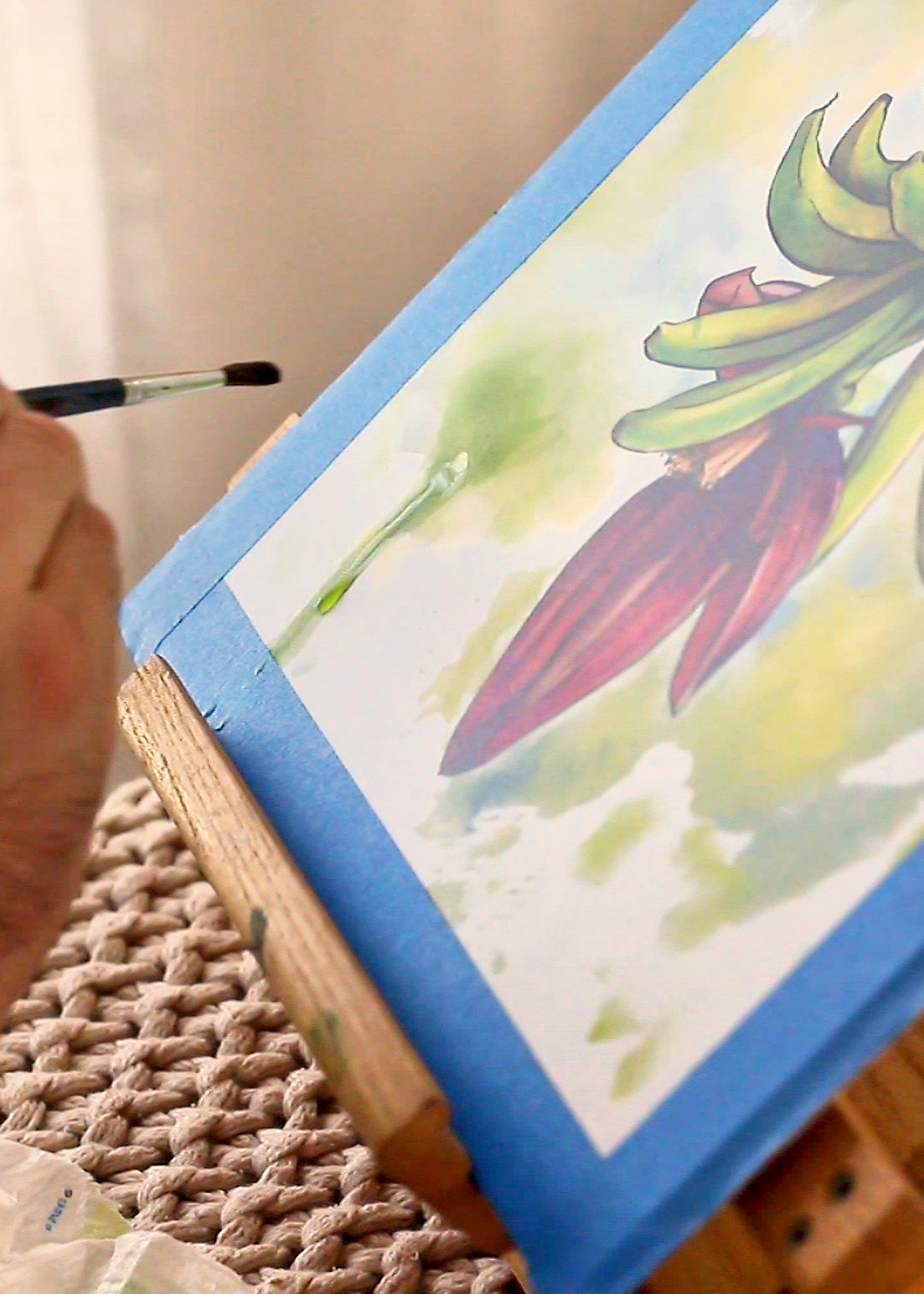A close-up of a hand holding a paintbrush and painting a watercolor illustration of a banana plant with green leaves and a purple-brown flower on a canvas taped to a wooden easel.