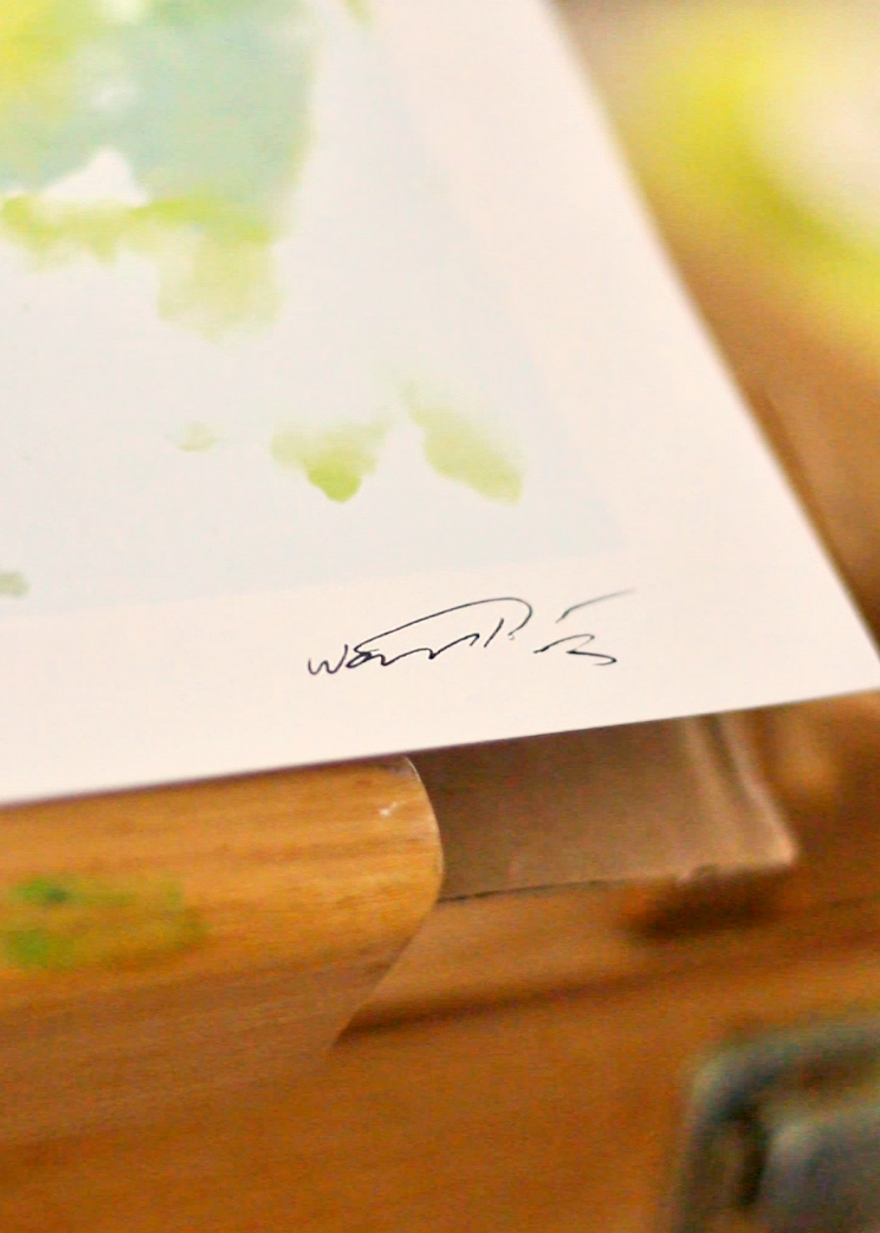 A close-up of a watercolor art print resting on a wooden easel or box. The print features soft green and yellow washes on white paper with a small black artist signature near the bottom.